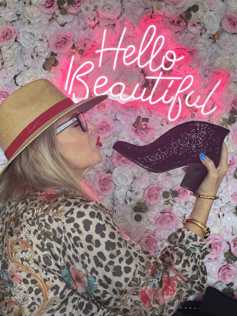 Woman with long hair, wearing a hat and a patterned blouse, kisses a glittery high-heeled shoe against a floral backdrop with “Hello Beautiful” neon sign.