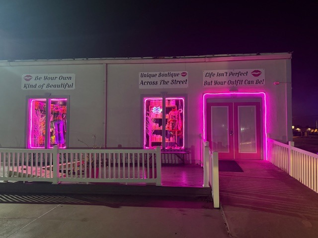 A brightly lit boutique exterior features neon pink accents and playful slogans promoting self-expression and fashion, inviting customers to explore.