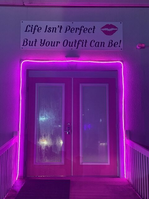 Bright pink doors framed by purple neon lights, topped with a sign reading "Life Isn't Perfect But Your Outfit Can Be!" signaling a trendy boutique.