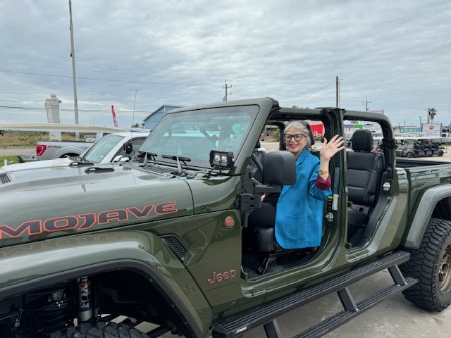 A woman in a blue jacket waves from the driver’s seat of a green Jeep Mojave at an outdoor event. The scene features cloudy skies.
