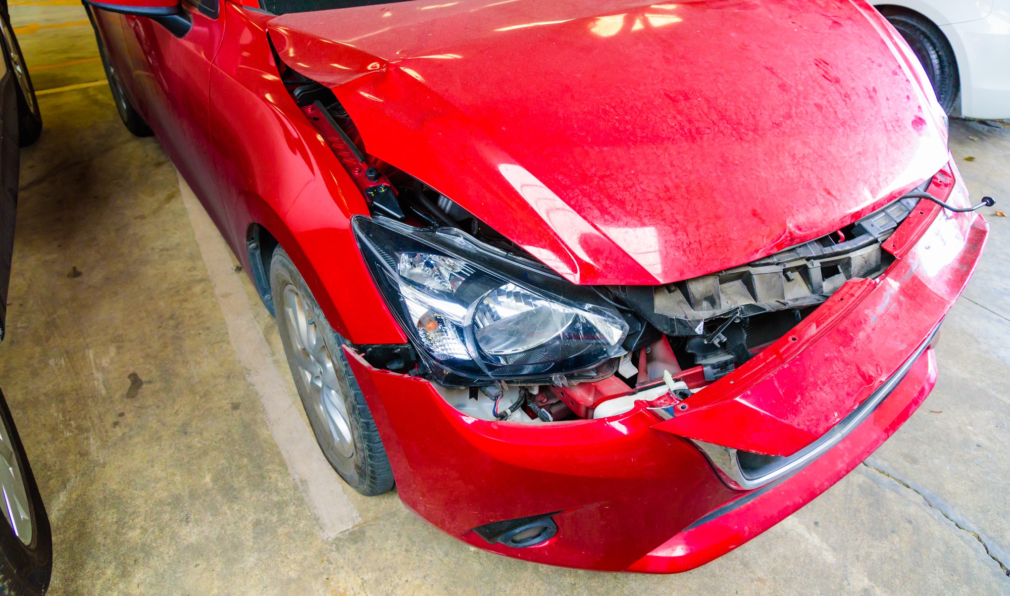 Damaged red car with a crumpled hood and exposed engine components, parked in a garage. Illustrates vehicle collisions and repair needs.