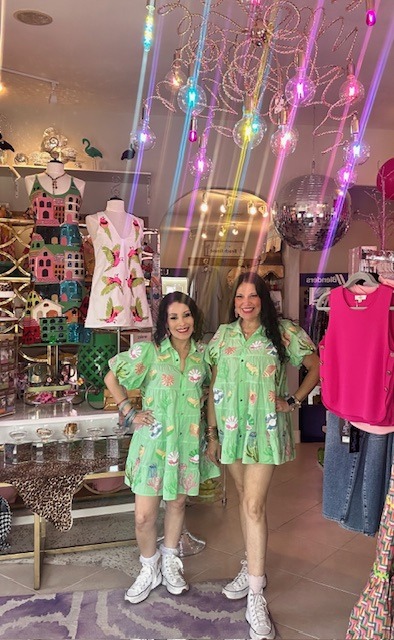 Two women stand in a colorful boutique, wearing matching green dresses with floral patterns. The shop features vibrant decor, including a disco ball and bright lighting.