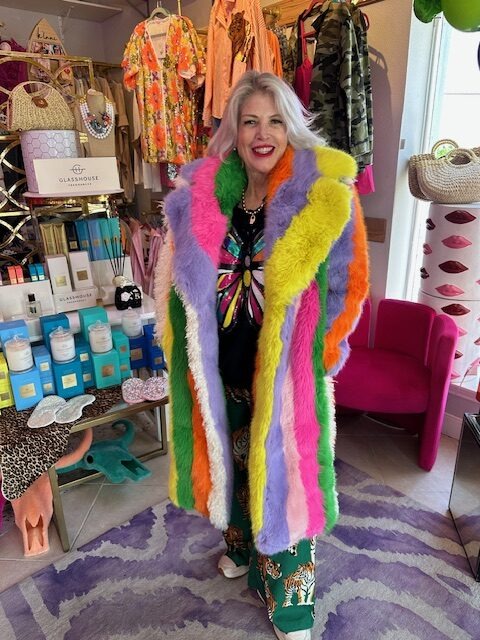 A woman in a vibrant, multicolored faux fur coat stands in a boutique filled with clothing and beauty products, showcasing bold fashion.