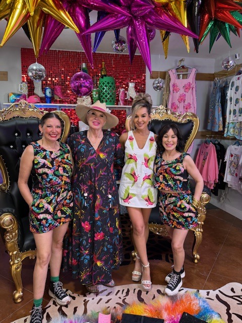 Four women in vibrant attire pose together in a boutique filled with colorful decorations and clothing. The setting exudes a fun, festive atmosphere.