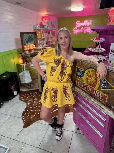 Woman in a yellow dress with tiger prints poses confidently beside a vintage gasoline sign in a vibrant, retro-themed café.