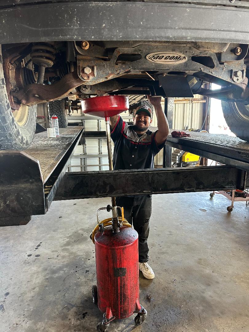 Mechanic working under a vehicle in a garage, holding an oil pan while collecting fluid. A red oil extractor is nearby, emphasizing vehicle maintenance.